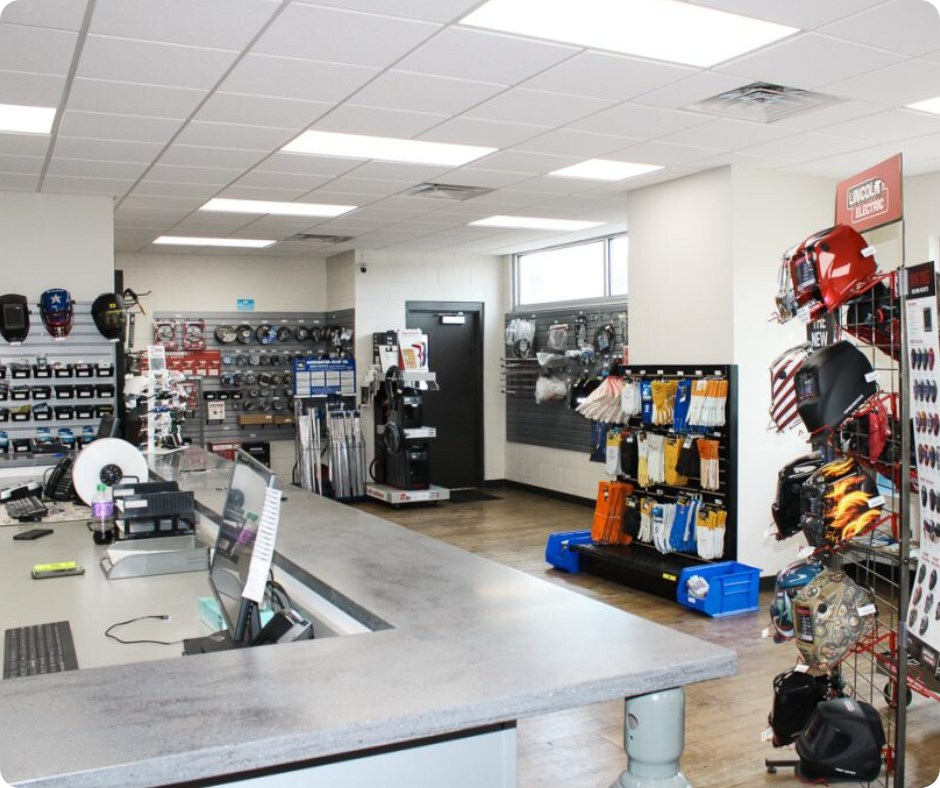 This photo showcases the retail counter and showroom of Metro Welding Supply Corporation in Detroit, MI. The store is well-lit and organized, featuring a wide range of welding equipment, tools, and safety gear.

Key Features in the Photo:
Front Counter Area:

A large, L-shaped sales counter with computers, phones, and office supplies, indicating a customer service and checkout area.
A clean and modern design with a polished gray countertop.
A point-of-sale system for processing orders.
Welding Helmets & Safety Gear Display:

Lincoln Electric welding helmets are prominently displayed on a red wire rack to the right.
Additional welding hoods and protective headgear are hung on the walls and shelves.
Shelving with Welding Supplies & Tools:

Various gloves, welding sleeves, and safety equipment are neatly organized on racks.
An assortment of grinding discs, welding rods, and consumables is stocked along the back wall.
A section of welding tools and accessories, including electrodes, cables, and connectors, is visible.
Rental & Equipment Section:

The background features industrial welding machines and rental tools, available for purchase or rent.
A black equipment stand houses additional welding-related tools and gear.
Clean & Professional Store Layout:

The store has a modern, organized layout, making it easy for customers to browse and find the equipment they need.
Bright LED lighting and white walls provide a clean, professional appearance.
A hardwood-style floor adds to the industrial yet welcoming feel.

This showroom exemplifies Metro Welding Supply Corporation’s commitment to providing high-quality welding equipment, gases, and safety gear for professionals and businesses in Southeast Michigan.