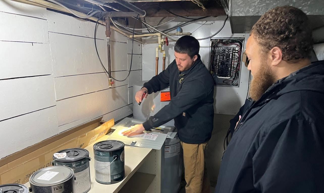 Home inspector shining a flashlight on an equipment panel door in a basement while a client watches.