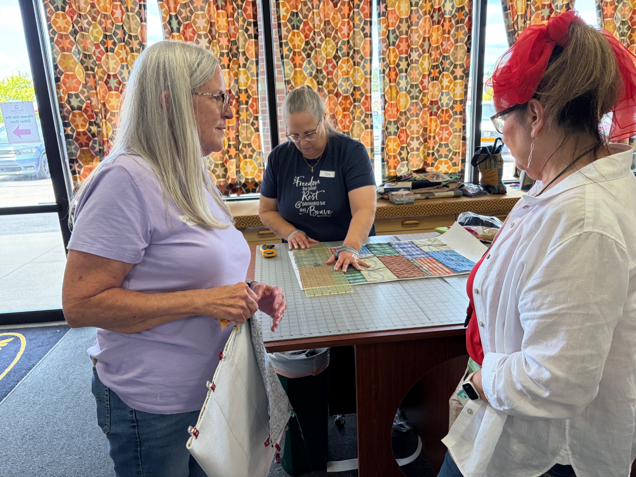 Students gather around for a fabric cutting demonstration during a Decorative Stitch class. Participants learn accurate measuring, rotary cutting, and piecing techniques to create precise quilting projects.