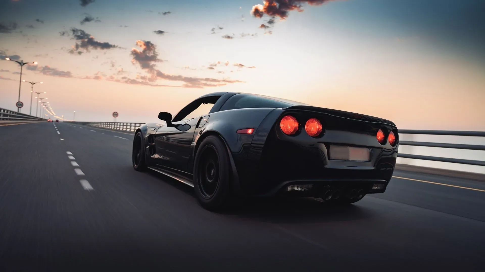 A black Chevrolet Corvette Z06 is captured in motion on a highway bridge during a sunset. The car's distinctive red taillights glow against the twilight sky, which features scattered clouds tinged with orange and yellow hues. Streetlights line the left side of the road, creating a sense of depth and perspective. The image conveys a feeling of speed and freedom.