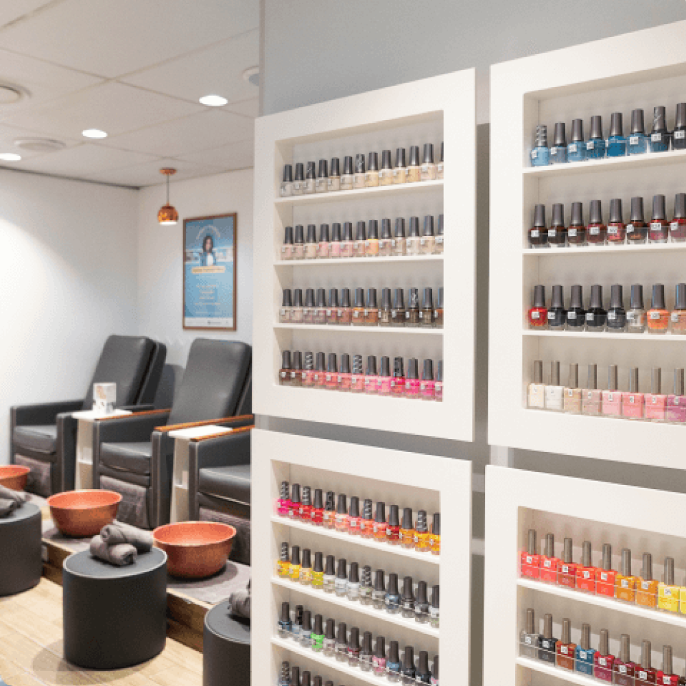 A modern nail salon interior with neatly arranged shelves displaying a wide range of nail polish colors. In the background, there are comfortable black pedicure chairs with orange foot bowls and rolled towels prepared for clients.