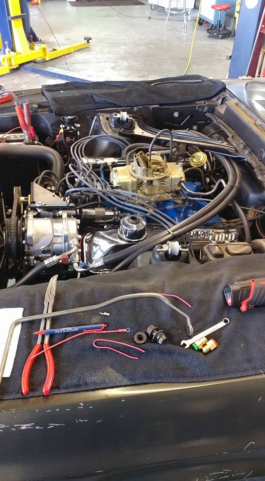 A close-up view of a car engine with various tools and parts laid out on a black cloth covering the engine bay. The engine appears to be undergoing maintenance or repair. Tools like pliers, a wrench, and electrical connectors are visible, alongside engine components like the carburetor and spark plug wires. The background suggests a workshop or garage environment with a car lift and industrial flooring.