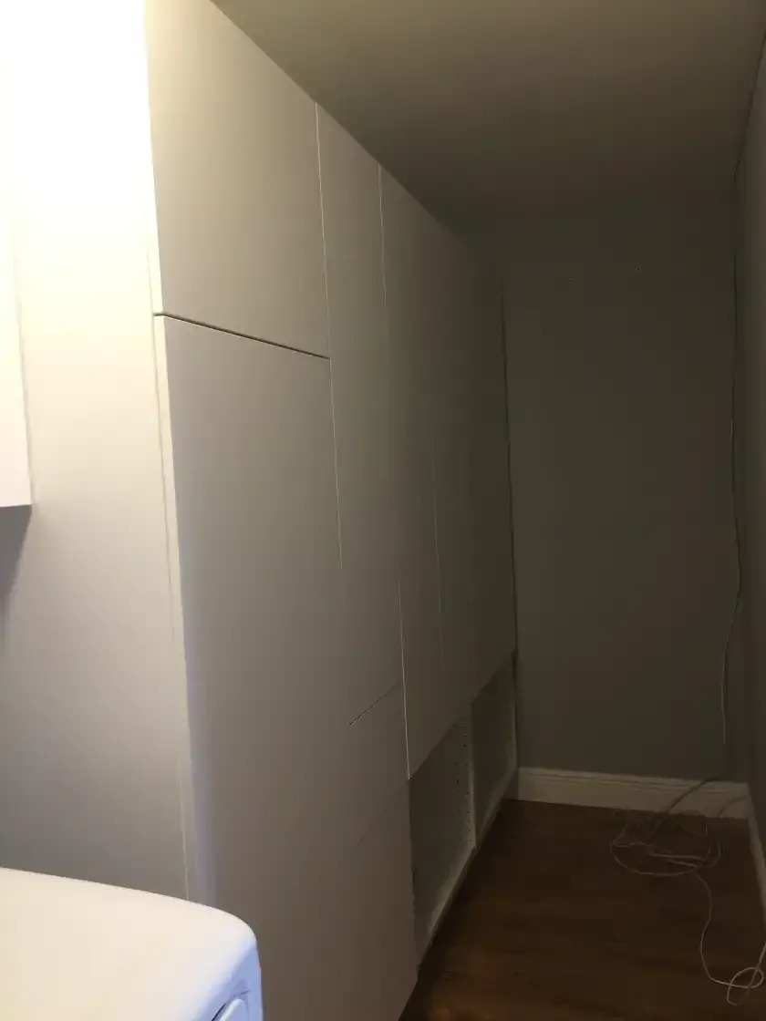 The image shows a white, built-in IKEA PAX wardrobe system in a room with wood flooring and gray walls. The wardrobe has multiple sections and drawers.