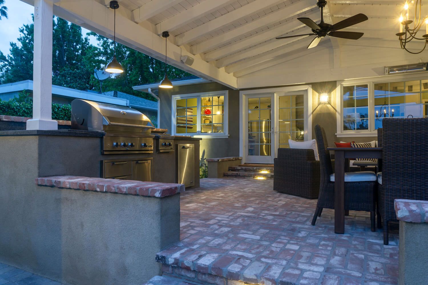 Covered outdoor patio with stainless-steel grill, brick flooring, dining table, ceiling fan, and warm lights creates an inviting retreat.