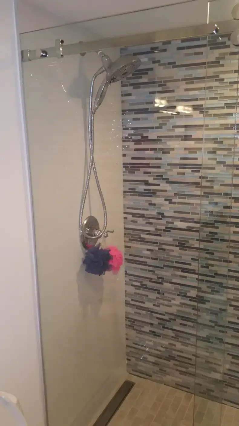 A modern bathroom features a glass-enclosed shower with horizontally striped gray and white tile. The shower head and faucet are silver, and blue and pink sponges hang inside.