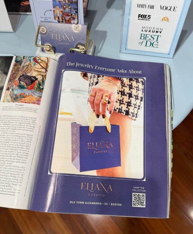 Eliana Curated&rsquo;s magazine advertisement features a purple shopping bag, statement ring and layered bracelets. The tabletop display pairs the jewelry campaign with its branded cards and media mentions.