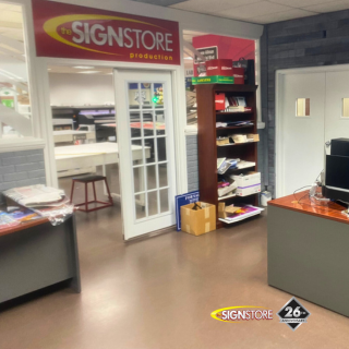 Signstore graphic designer's office with a desk displaying large letters and various papers. The office has a view of the production area through a glass window. The "26" anniversary badge is visible.