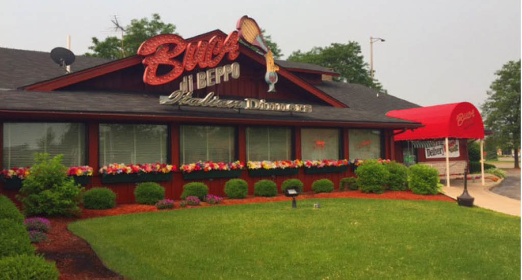 Buca di Beppo Lombard shows a big Buca sign, red walls, flowers along the windows, and greenery.