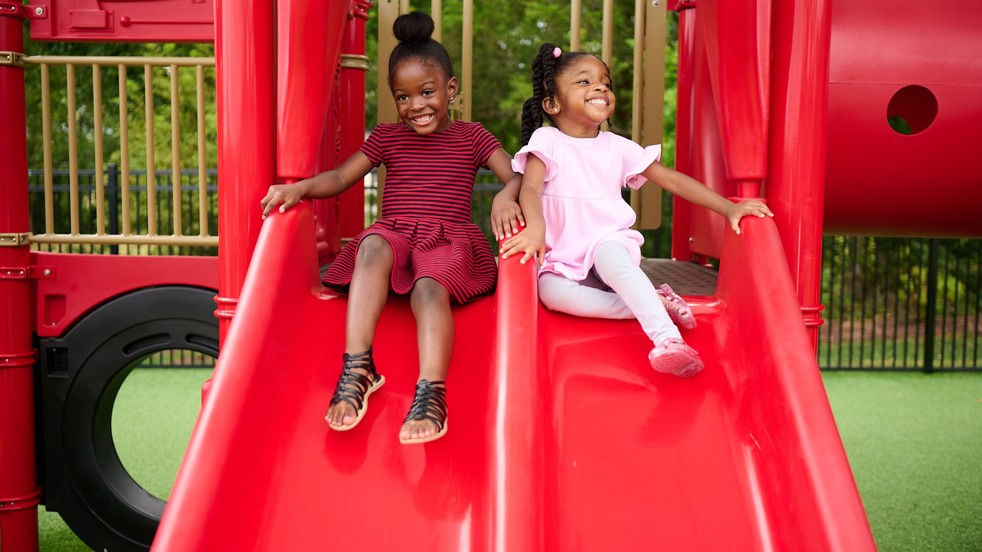 Two Primrose Schools children smiling together on red playground slide during outdoor play time