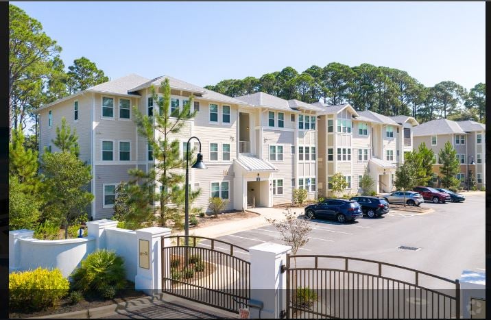 A gated entrance leads into a modern three-story apartment community nestled among lush pine trees.