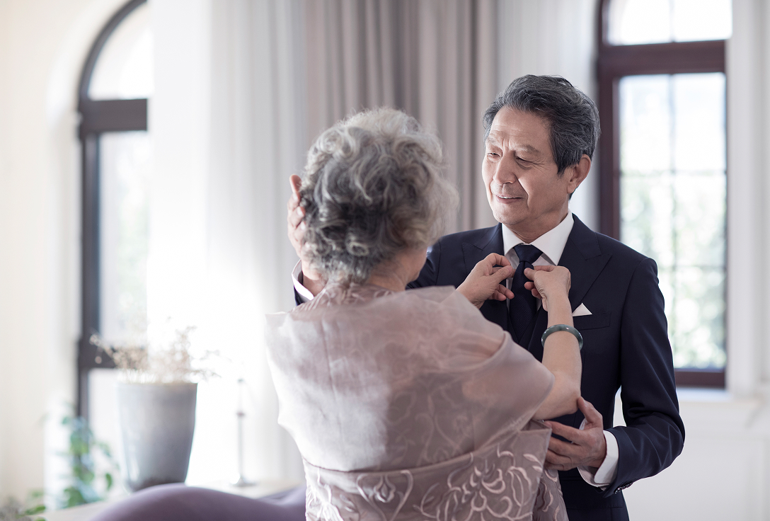 Elderly couple in a softly lit room; woman adjusts man's tie affectionately. Both are elegantly dressed, conveying warmth and love.
