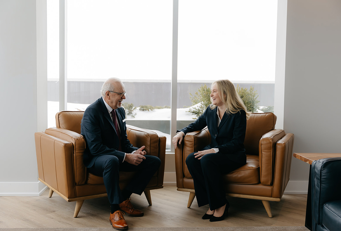 Two people in formal attire sit in brown leather armchairs, engaging in a warm conversation near a large window. The setting is modern and professional.