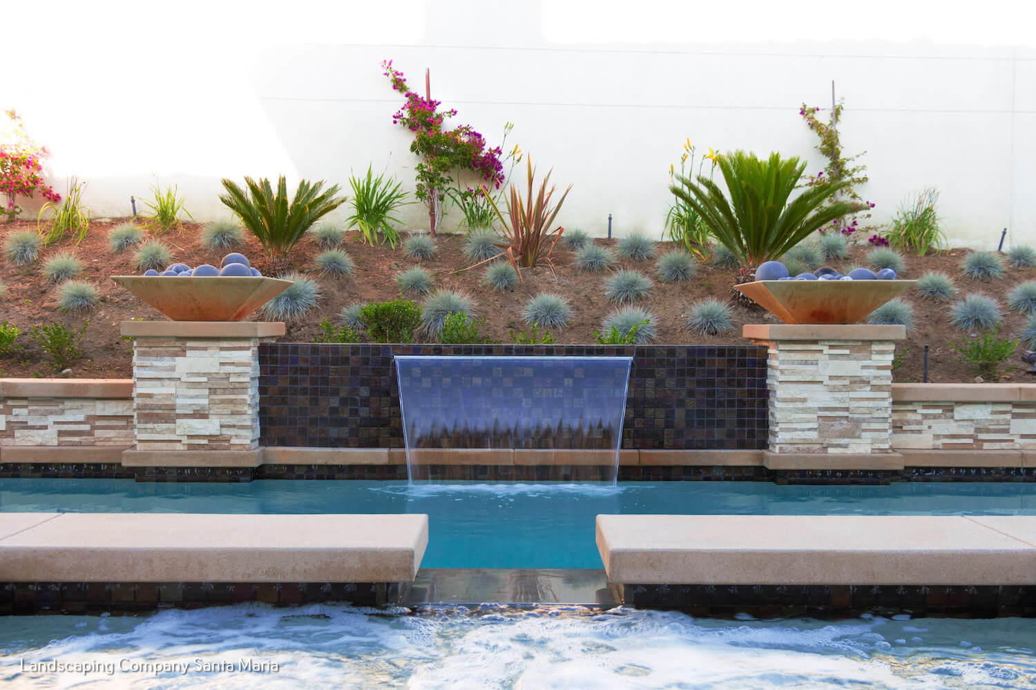 Modern backyard pool with a blue-tiled waterfall, raised stone planters, succulents, palms, and colorful flowers creating a serene resort-like landscape.