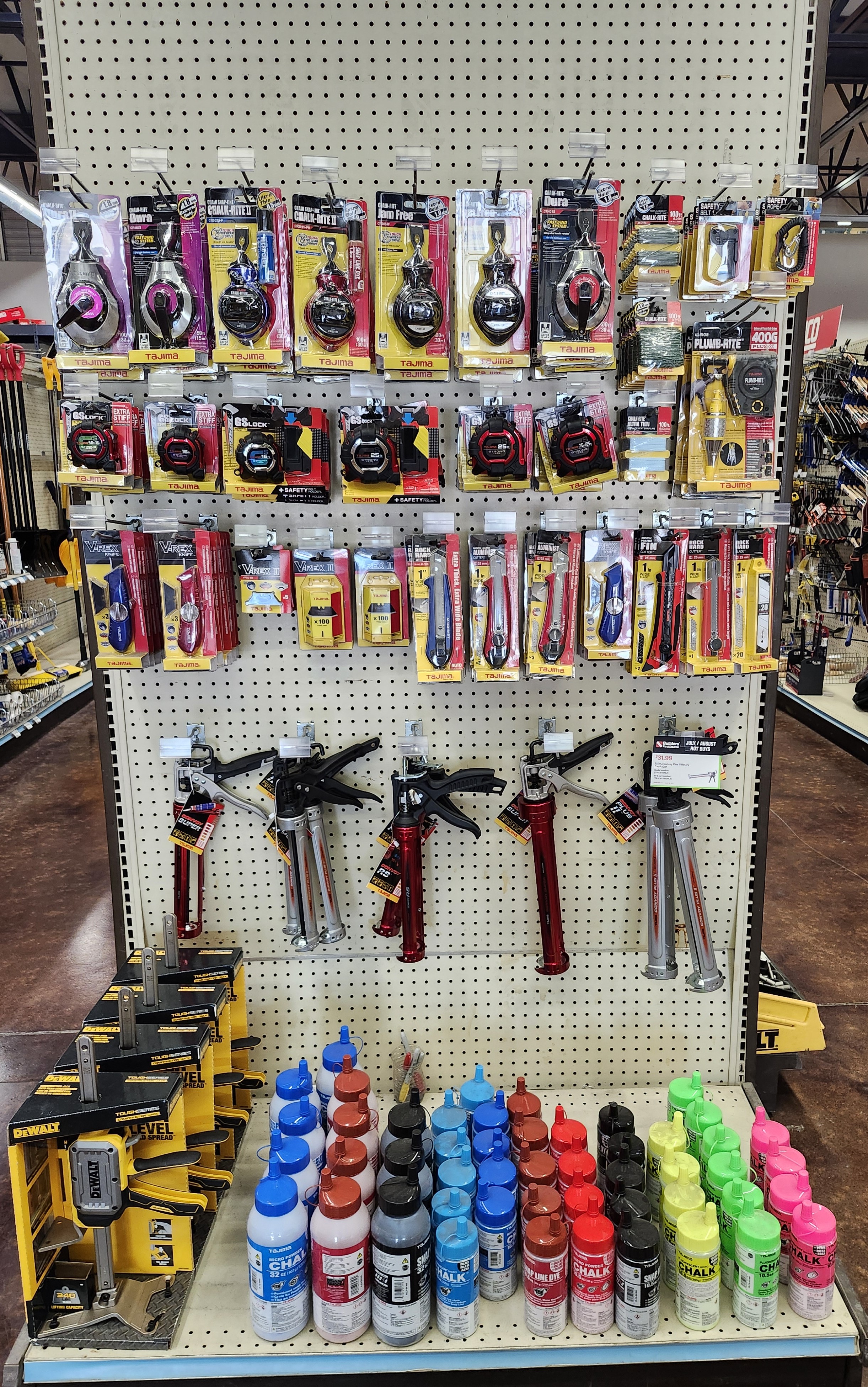 A well-organized display of various Tajima products including chalk lines, measuring tapes, utility knives, caulking guns, and colorful bottles of chalk at a Builders FirstSource lumber yard in Midvale, UT. The tools are neatly arranged on a pegboard with clearly visible branding and packaging.