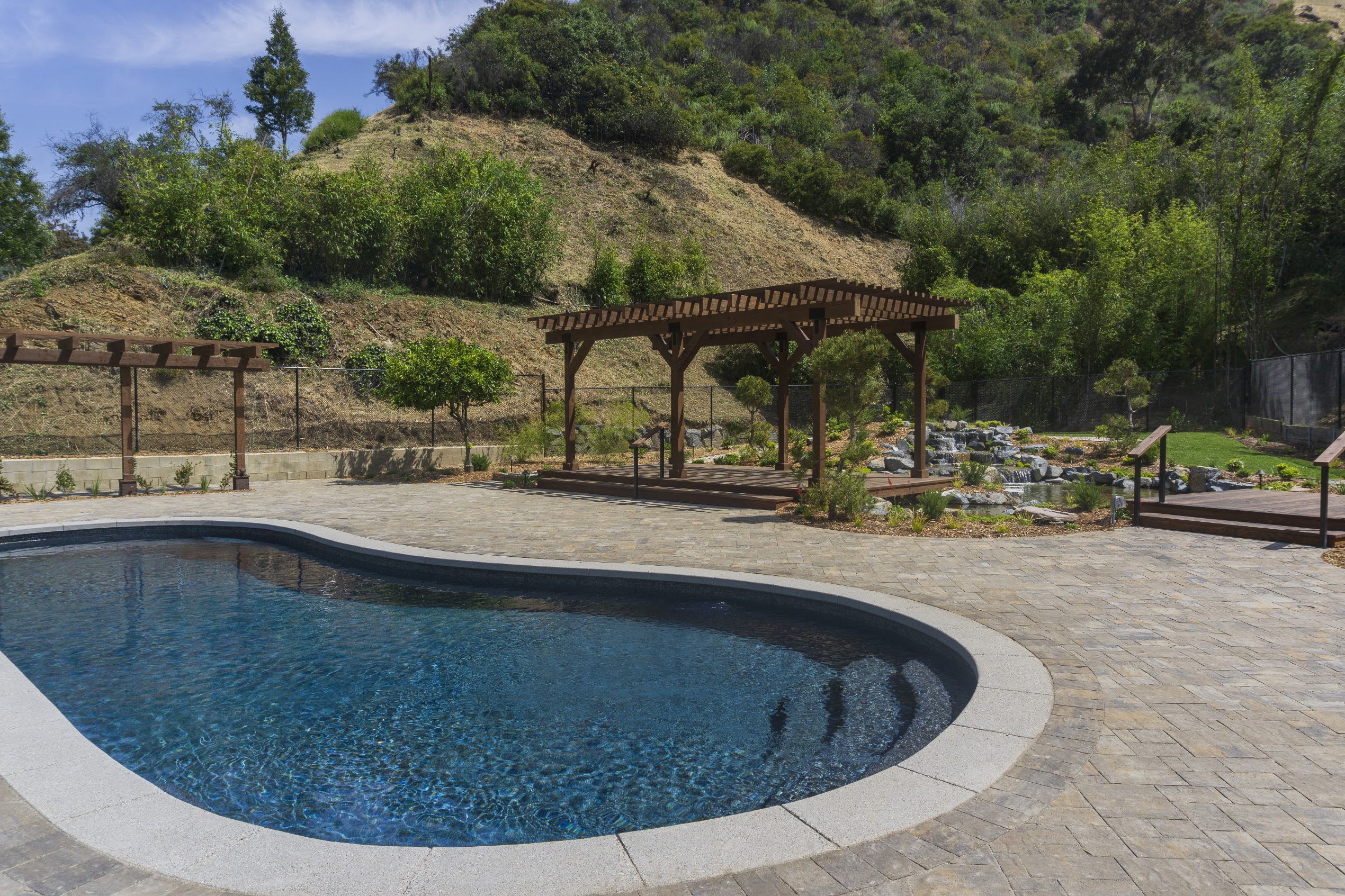 Curved backyard swimming pool beside a rustic wooden pergola, landscaped hillside, stone patio, and sunny blue sky create a serene retreat.