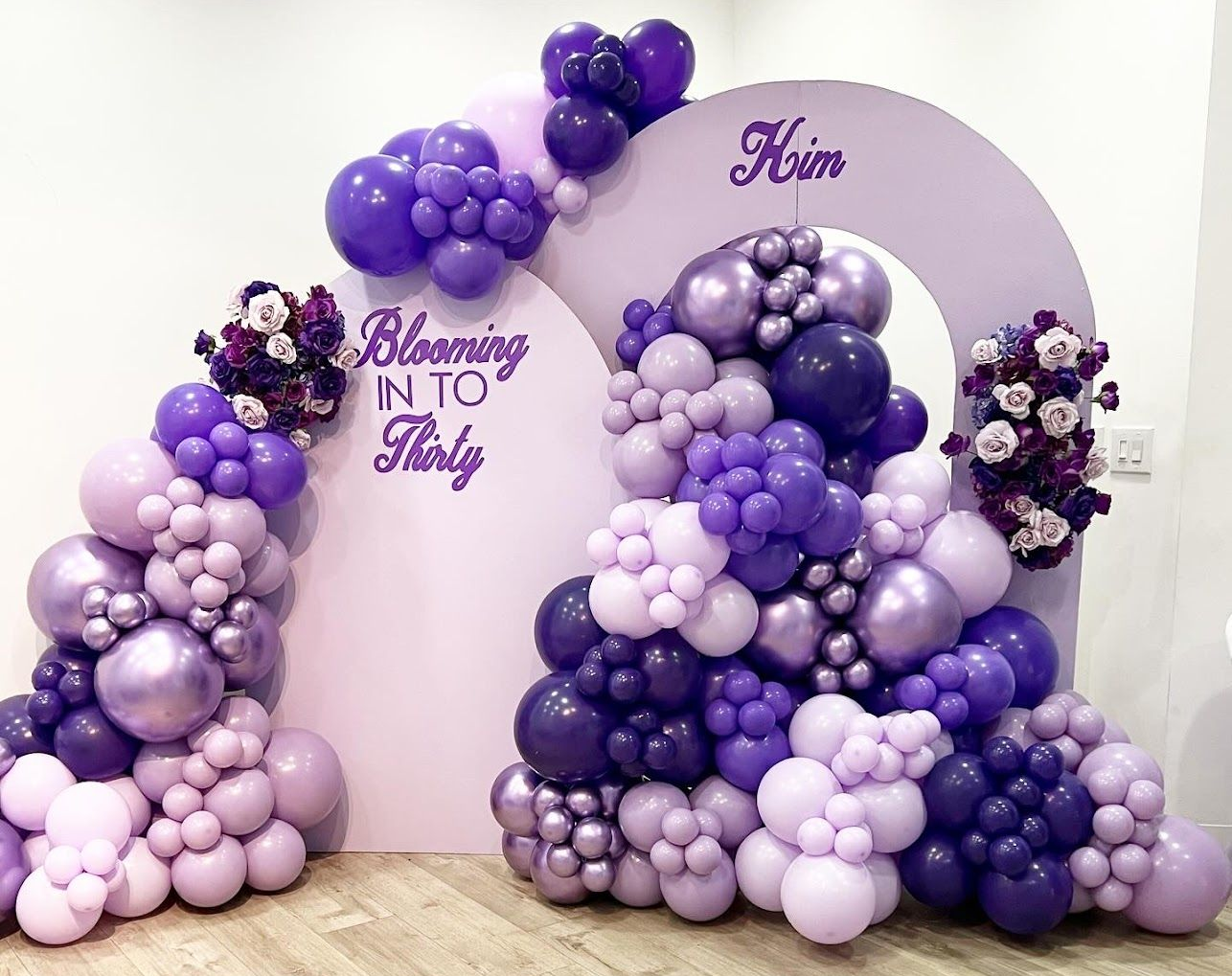 Elegant milestone birthday backdrop featuring purple and lavender balloon garlands, floral accents, arched panels with Blooming Into Thirty text, and Him name, creating a stylish, celebratory indoor photo display setting.