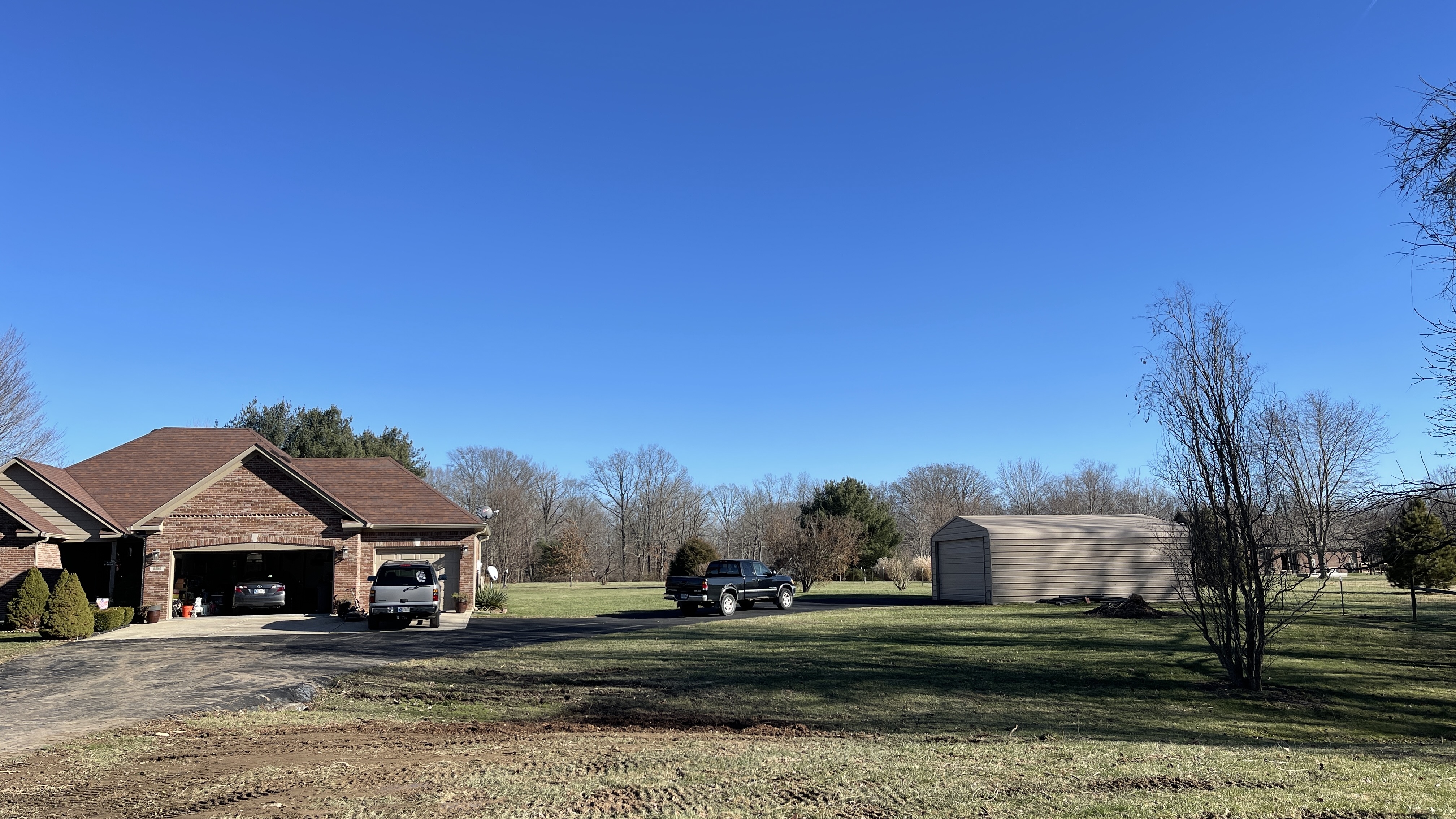 A suburban landscape under a clear blue sky, featuring a brick house with a garage, a separate metal shed, two pickup trucks, and several bare trees. The foreground shows a patchy lawn with visible tire tracks, suggesting recent activity or landscaping. The overall scene depicts a residential property on a sunny day.