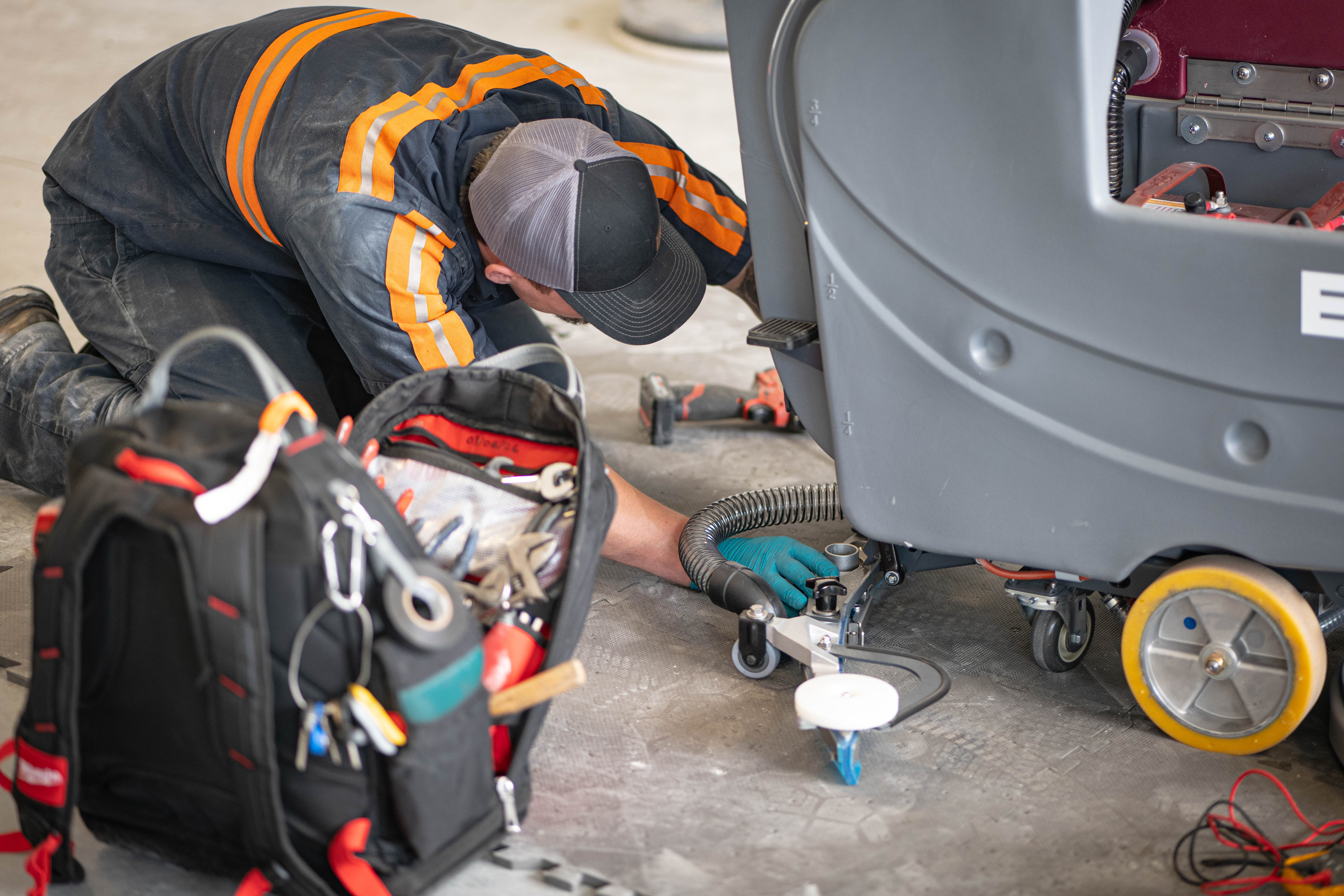 A low-angle view of a WaySolutions technician inspecting the lower vacuum hose connection and caster wheel mechanics on a grey Minuteman E20 automatic floor scrubber. A well-stocked Milwaukee tool backpack sits open in the foreground, showcasing on-site mechanical repair readiness for industrial and supermarket cleaning systems.