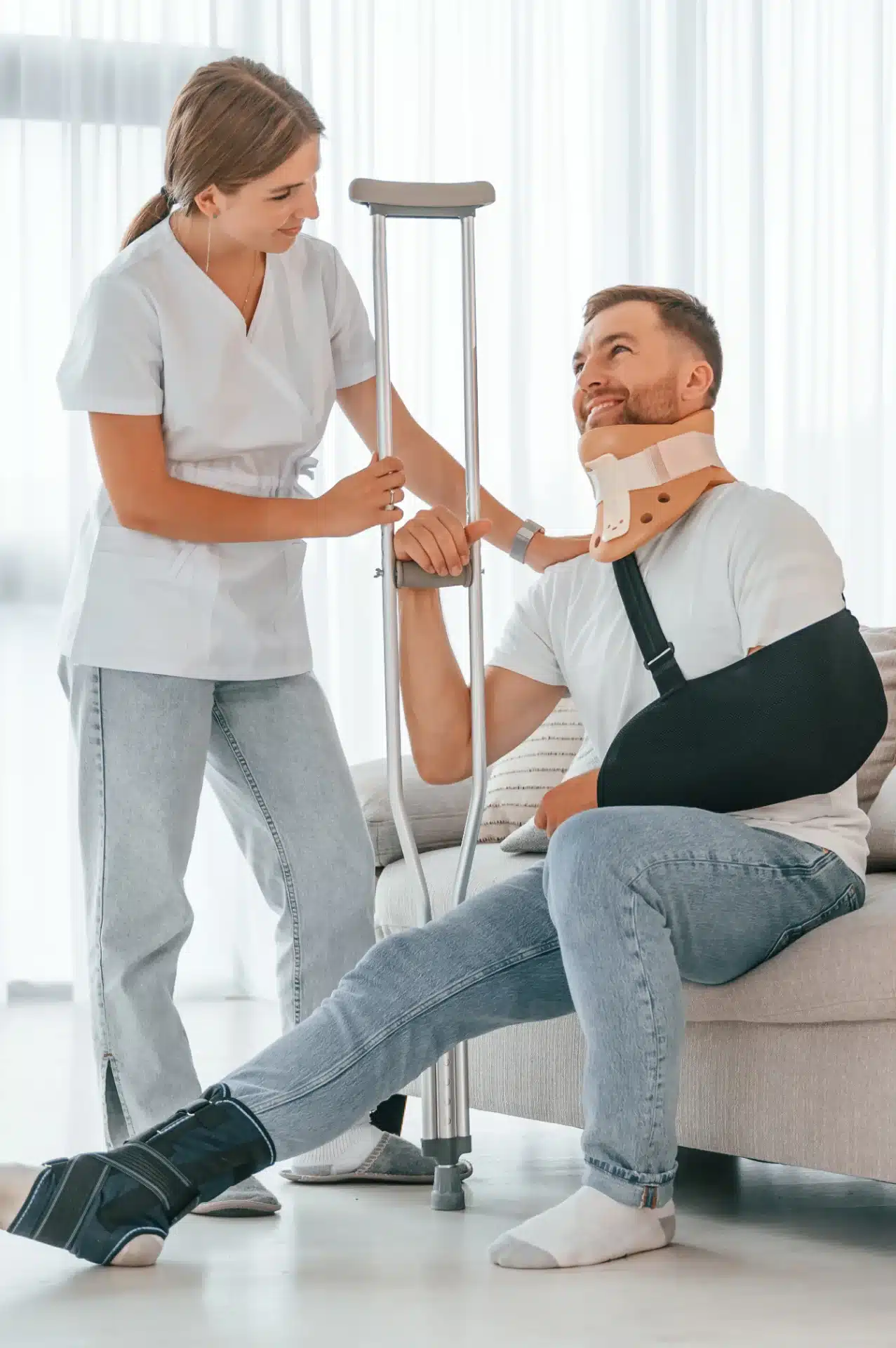 A woman in a white medical uniform assists a man with a neck brace and arm sling, who is seated on a couch and has crutches. The man is also wearing an ankle brace. The scene appears to be a home or clinic setting with bright natural light. The woman is holding onto the crutches, and they are both smiling, suggesting a supportive interaction.