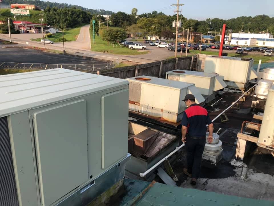 HVAC technician inspecting rooftop air conditioning units on commercial building, walking between systems and ductwork, with surrounding street, vehicles, and businesses visible, demonstrating routine maintenance and professional service work environment.