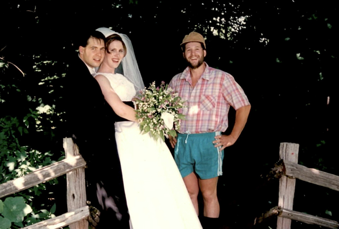 A bride and groom in formal attire, smiling at the camera with a casually dressed man in shorts on a wooden bridge in a lush, green outdoor setting.