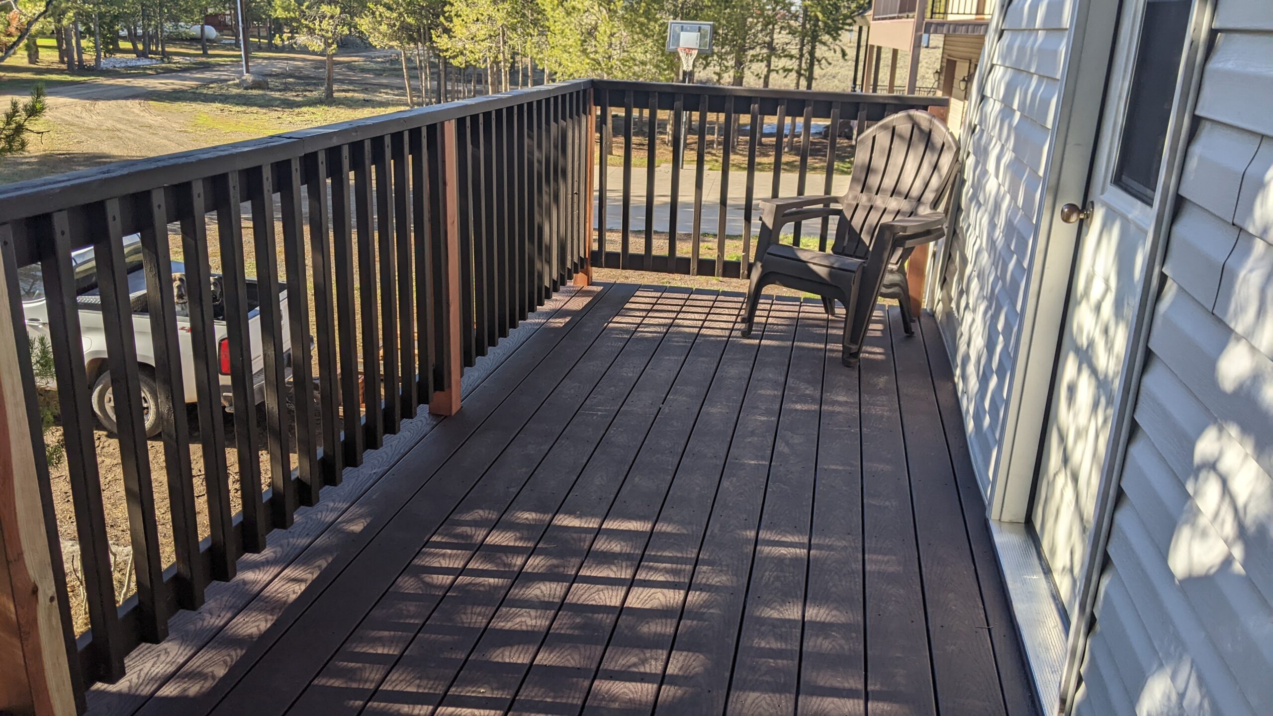 Extend your upper-level living space with this cozy raised composite deck by DeckPro LLC, designed for tranquility and function. We installed low-maintenance composite decking in a warm brown tone and a matching perimeter railing system. Furnished with a comfortable dark Adirondack chair, it's the perfect retreat to take in the serene views of the surrounding pine trees and rustic outdoor setting.