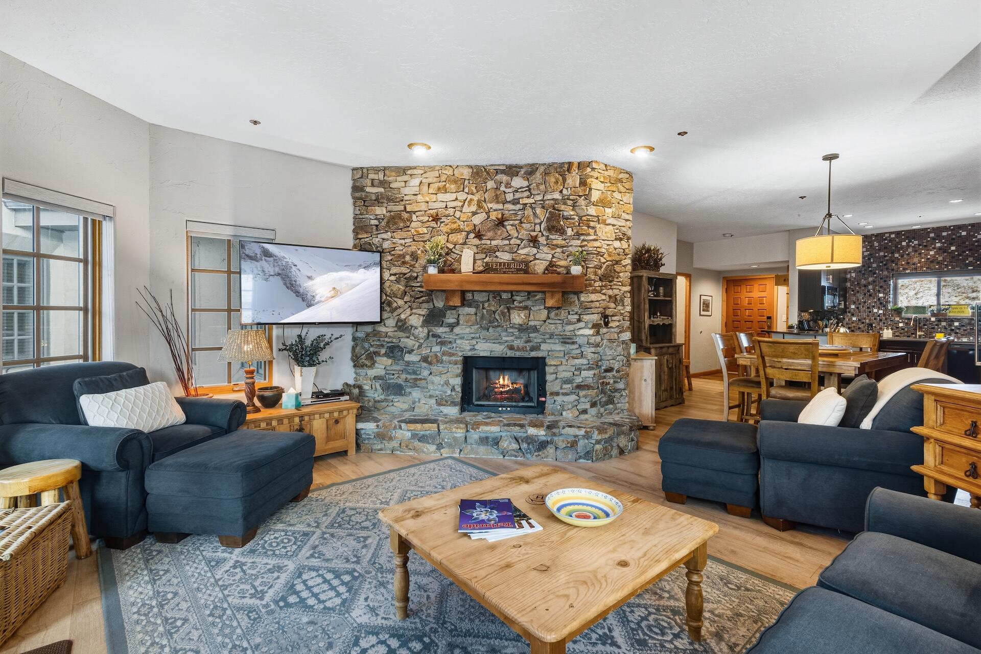 Condominium living room with blue seating, a stone fireplace, and an adjoining dining area.