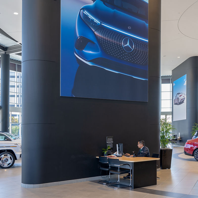 A modern, spacious car dealership showroom with large digital advertisements for Mercedes-Benz vehicles. A man is seated at a reception desk, attending to a computer. Cars are visible in the background, along with large windows and interior plants, creating a clean and professional environment.