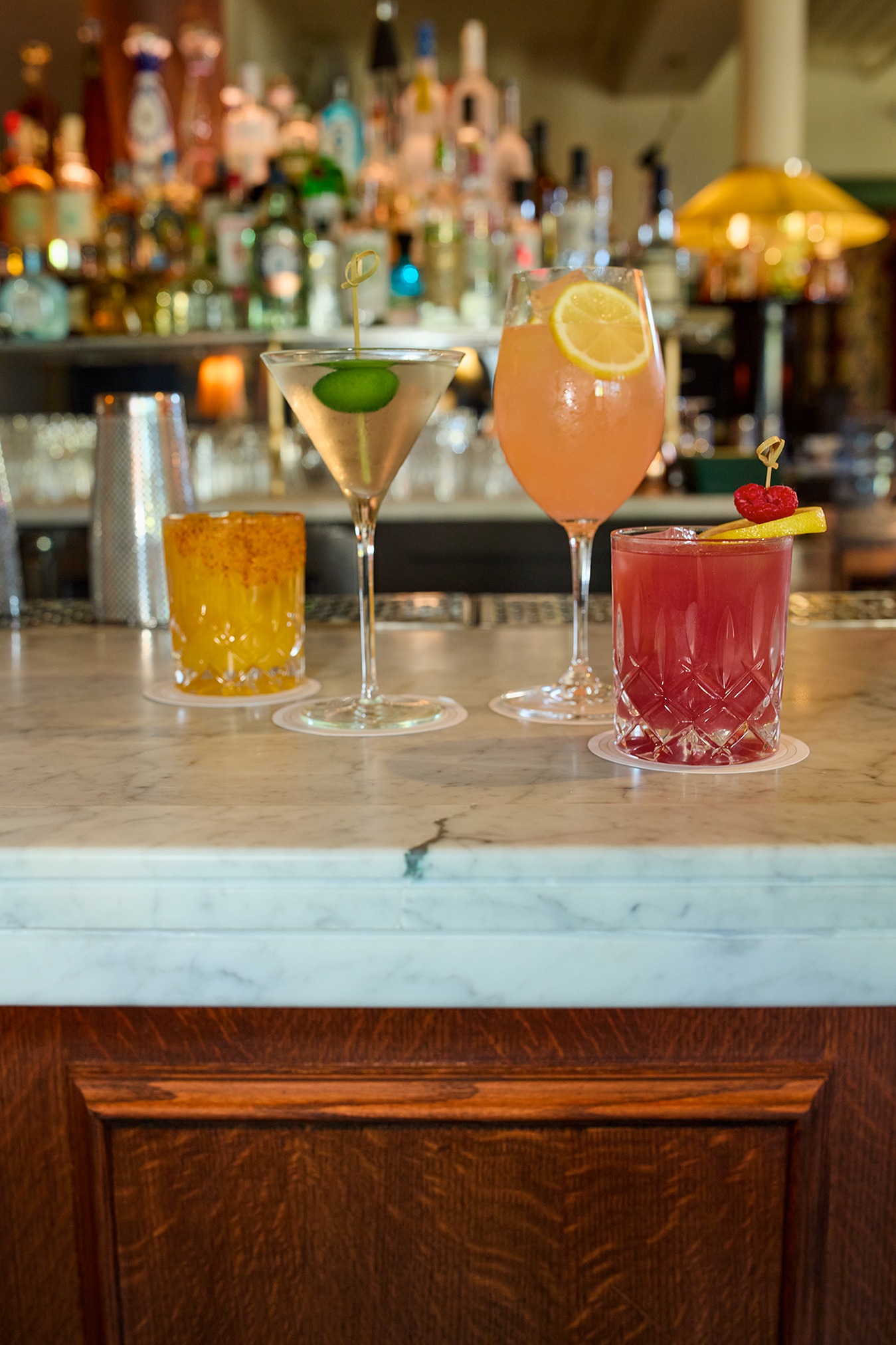 An array of drinks including a martini, a pink spritz with lemon, and two lowball cocktails in textured glassware. They are lined up on a white marble bar with a wooden base and soft background lighting.