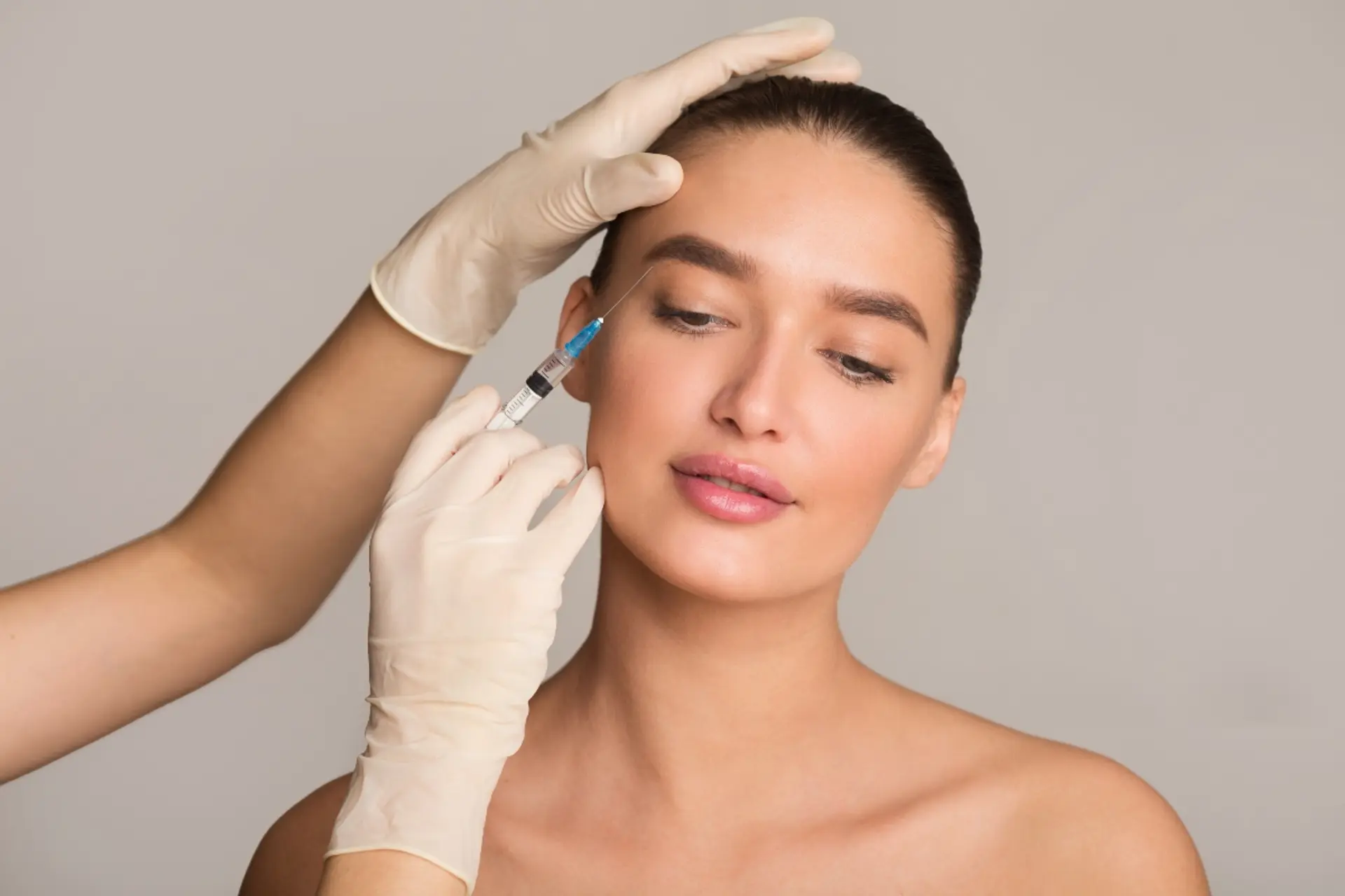 A close-up studio shot shows a person receiving an injection in the face. A medical professional's hands, wearing light beige disposable gloves, are visible holding a syringe and gently placing the needle near the person's temple. The person receiving the injection has their eyes looking down and slightly to the side, with their lips slightly parted. Their hair is pulled back, revealing their smooth skin. The background is a plain, neutral grey, emphasizing the subject and the medical procedure.