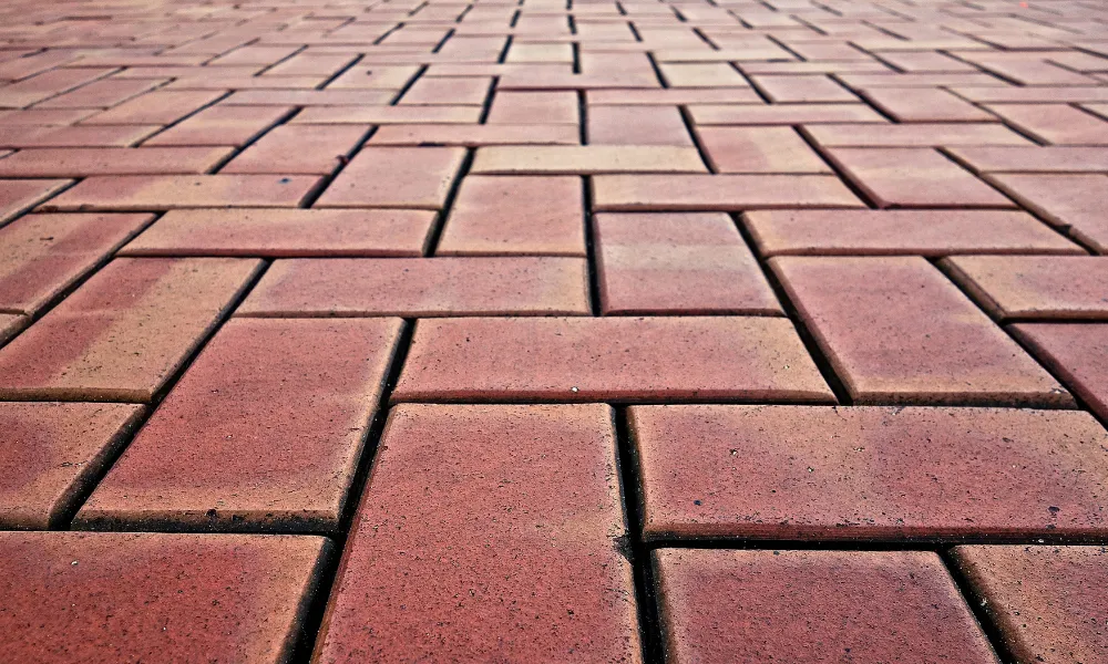 A low-angle, close-up shot of a red brick pathway laid in a herringbone pattern. The bricks appear textured and weathered, with variations in color from light reddish-orange to deeper red hues. The perspective draws the eye towards the vanishing point in the distance, emphasizing the repeating pattern of the interlocking bricks.