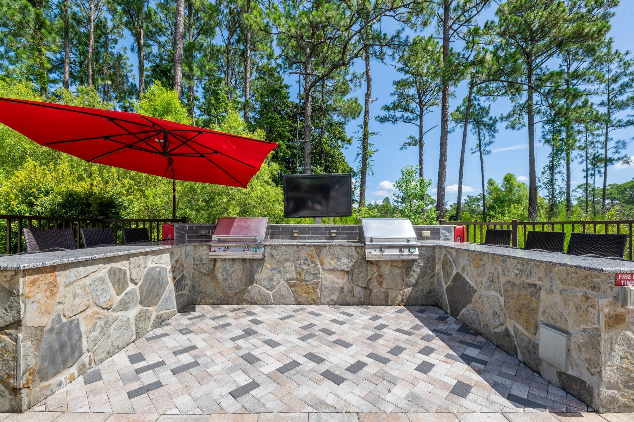 A premium outdoor grilling station features stone masonry, dual stainless steel grills, and a large red sun umbrella.