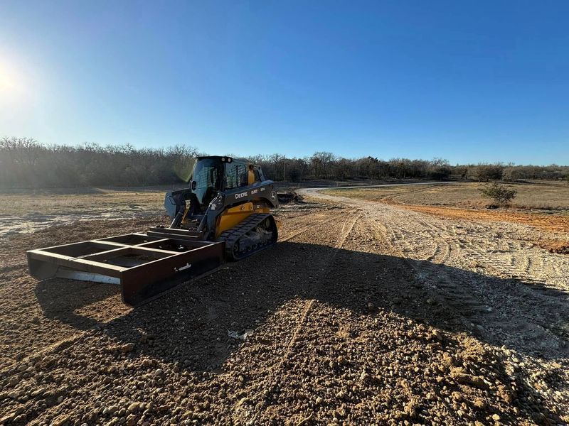 Skid steer grading large open land area with attachment, smoothing soil and preparing surface for construction, landscaping, or development, emphasizing precision grading and land leveling services in wide outdoor terrain.