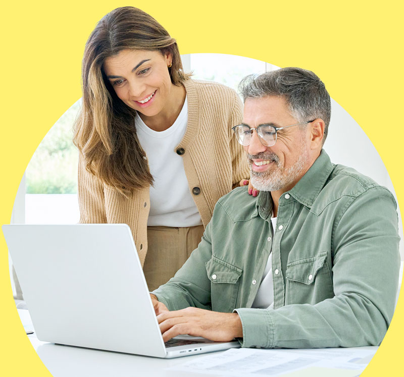 A man and a woman are looking at a laptop screen and smiling. The man is wearing glasses and a green shirt, and the woman is wearing a tan cardigan. They are both sitting at a desk with papers on it. The background is yellow and shows a blurred outdoor scene with trees and a window. They appear to be collaborating or sharing information.