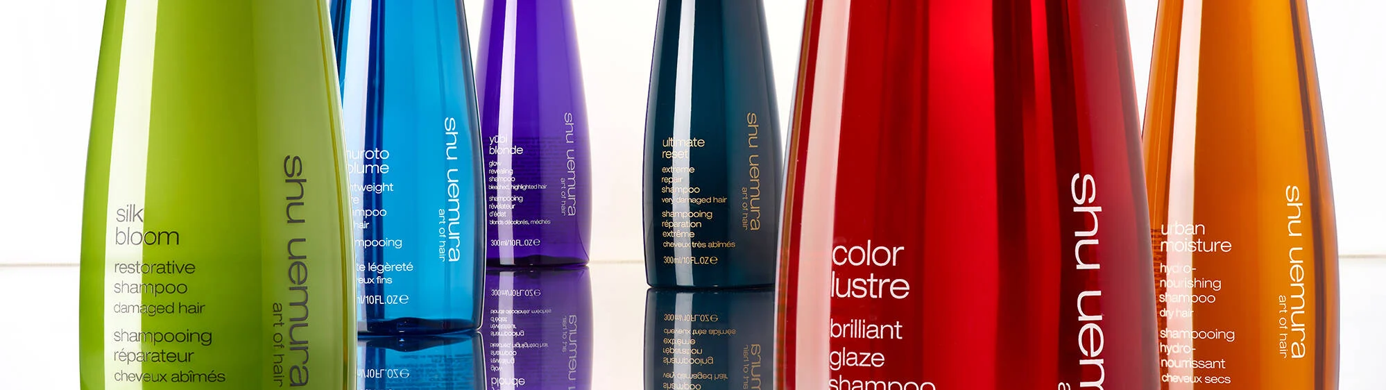 A collection of Shu Uemura hair products, featuring bottles in vibrant colors like green, blue, purple, red, and orange. Each bottle has white text detailing the product name and function. The bottles are arranged in a row and are reflected on a glossy surface below them.