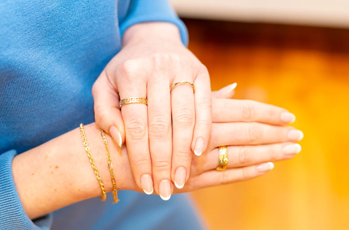 Gold-tone rings and delicate chain bracelets from Eliana Curated add a polished touch to a blue sweater. Explore stylish jewelry and everyday accessories in Old Town Alexandria, Virginia, for outings.