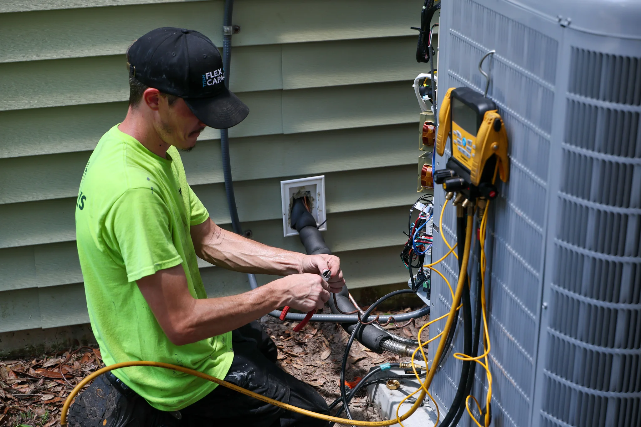 A man wearing a green shirt and a black baseball cap is kneeling in front of an outdoor air conditio 