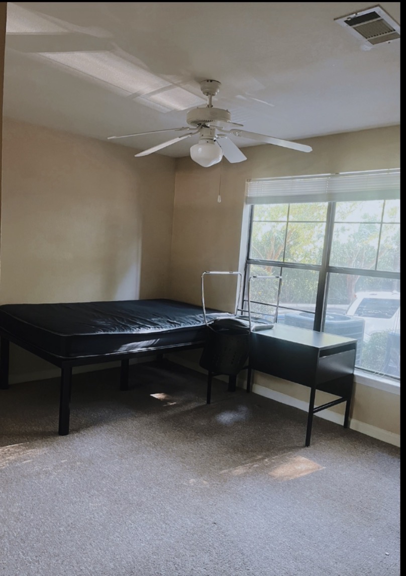A sparsely furnished room with a bed, a desk, and a chair next to a large window. The room has carpeted floors and beige walls. A ceiling fan is visible above.