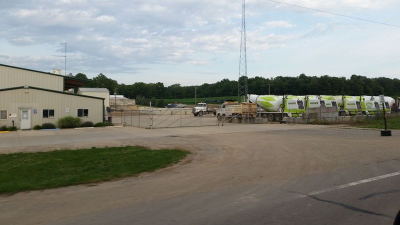 A wide view of Dave&rsquo;s Concrete Products facility shows multiple lime-green and white mixer trucks parked in a gated lot beside an office building, with a tall communication tower nearby.