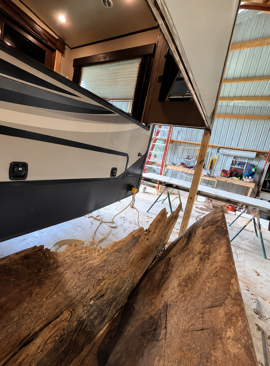 RV exterior under repair inside a workshop, showing an extended slide-out section and several removed, severely deteriorated wooden structural boards placed nearby, with ladders, tools, and workbenches clearly visible around.
