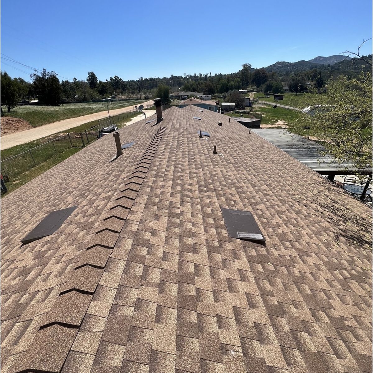 Overhead view down the central ridge of a wide brown shingle roof, showing vents and exhaust pipes, overlooking a rural landscape with open fields and hills under clear skies.