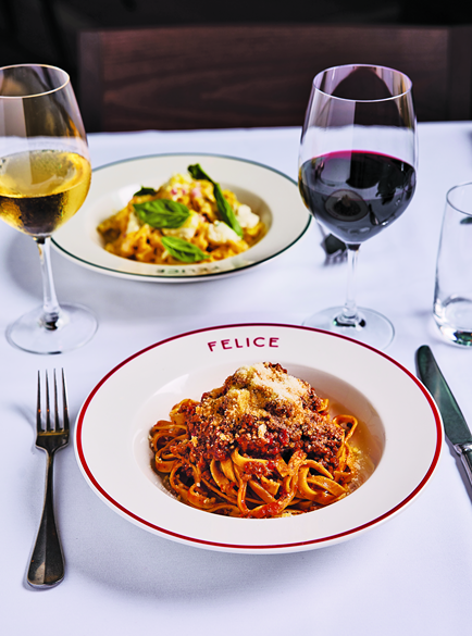 A plate of pappardelle bolognese sits in the foreground, flanked by a glass of red wine and a glass of white wine. In the background, another pasta dish completes the setting.