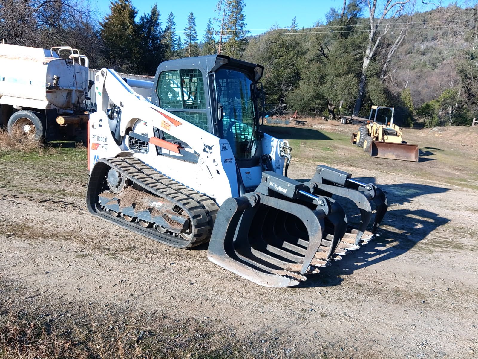 Tracked skid-steer loader equipped with a heavy grapple bucket sits on a dirt worksite, with a water truck and grader visible nearby among trees, hills, and clear blue sky surroundings.