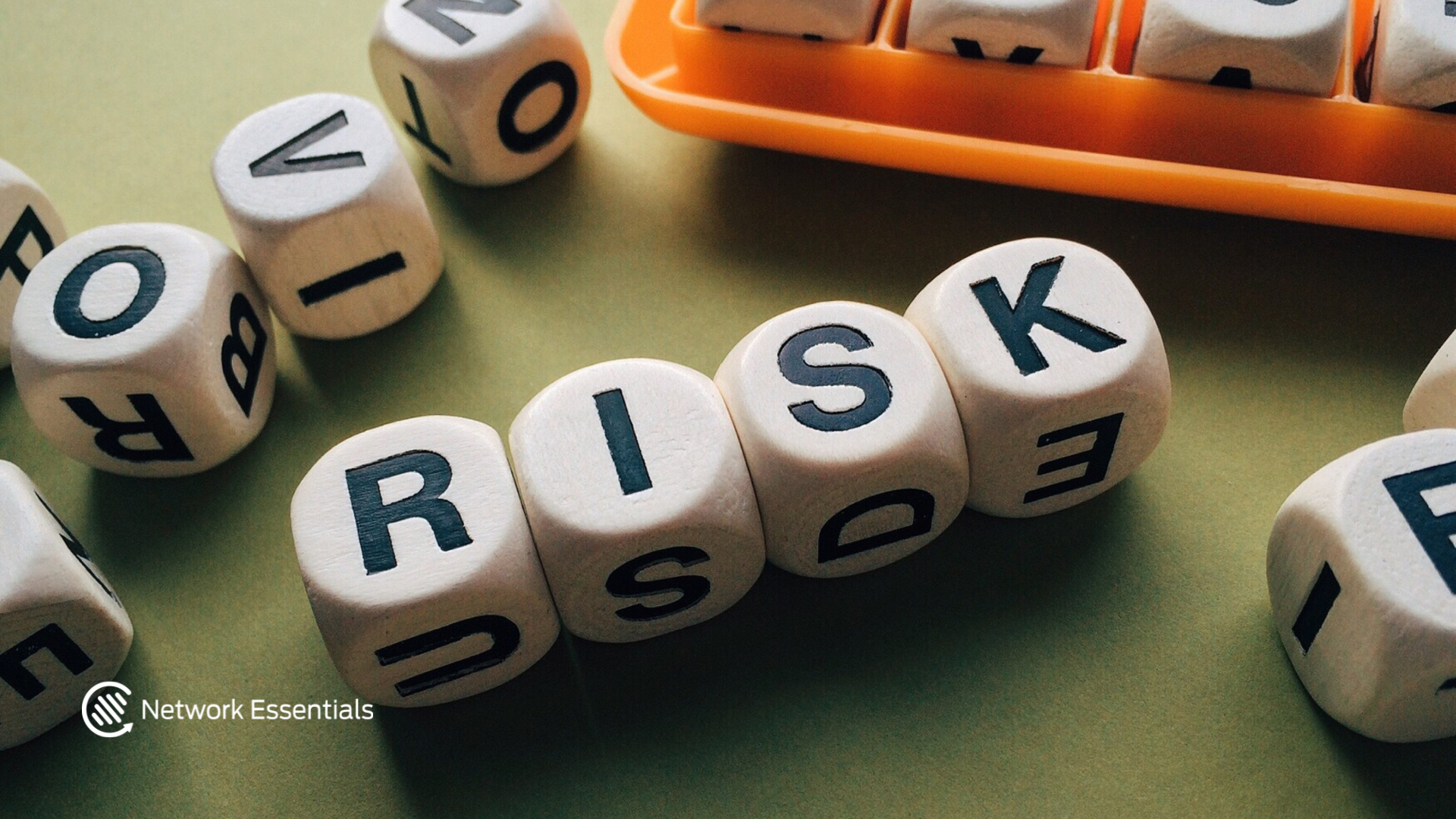 A close-up shot of wooden letter blocks spelling out the word "RISK" are arranged on a flat, olive-green surface. Other blocks with various letters are scattered around. In the background, an orange plastic tray holds more letter blocks. A "Network Essentials" logo is visible in the bottom left corner.