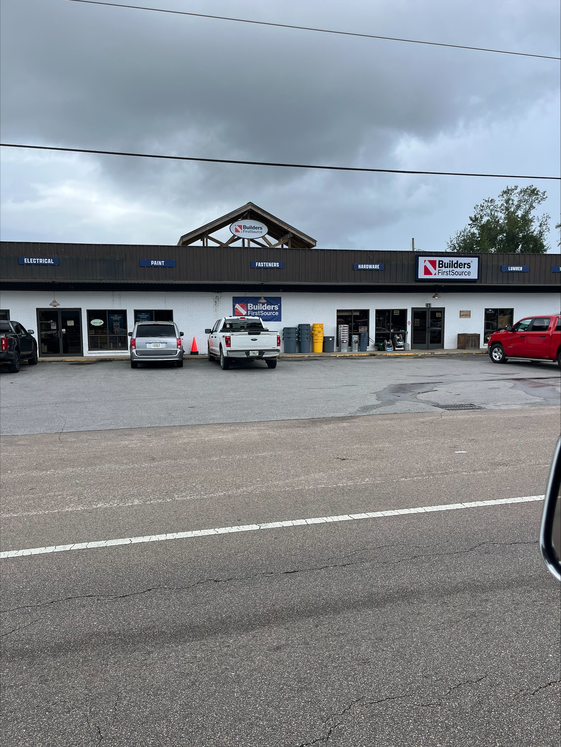 The image shows the exterior of a "Builders FirstSource" hardware store located at 244 FL-20, Freeport, FL. The building is a single-story structure with a dark brown and white color scheme. The store's name, "Builders FirstSource," is prominently displayed on two signs: one on the upper front wall and another above the entrance.

The upper front wall also features labels indicating various product categories:
Electrical, Paint, Fasteners, Hardware, Lumber

The entrance has multiple glass doors, and there are several items on display outside, including metal trash cans and other hardware supplies. The parking area in front of the store has a few vehicles parked, including a white pickup truck and a red truck. The sky is overcast, with dark clouds indicating possible rain. The road in front of the store has visible lane markings and cracks in the pavement.