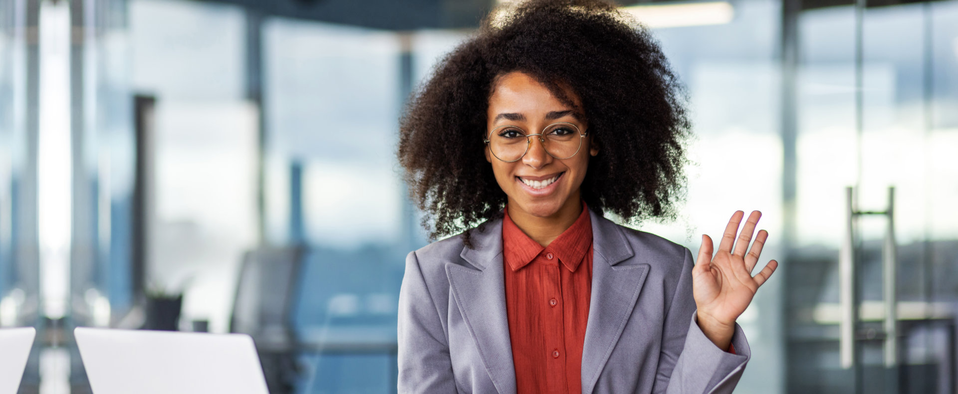 A smiling Black woman with curly hair and round glasses wears a grey blazer over an orange shirt. Sh 