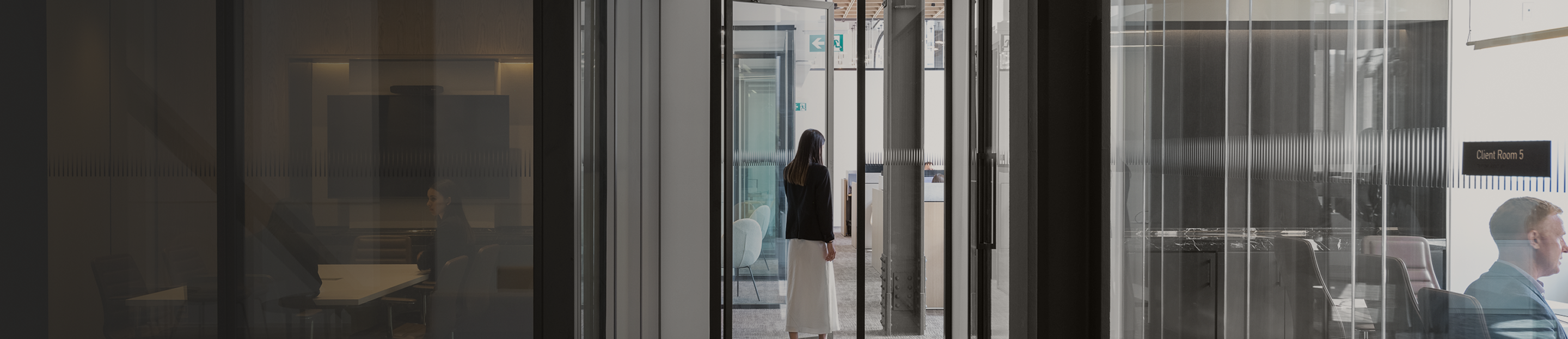Woman walking down a hallway with glass-walled boardrooms on either side.