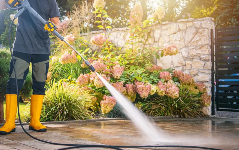 Professional using a pressure washer to clean a paved outdoor surface near a garden and stone wall. Wearing protective boots and gloves, the worker removes dirt and stains, restoring the patio&rsquo;s appearance while enhancing cleanliness and curb appeal.