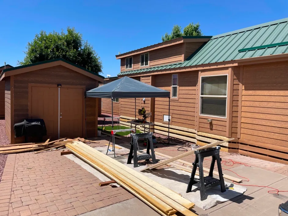 Residential exterior painting preparation scene with wooden boards, sawhorses, and tools set up beside a brown house and shed, showcasing an active workspace for renovation under clear blue skies.