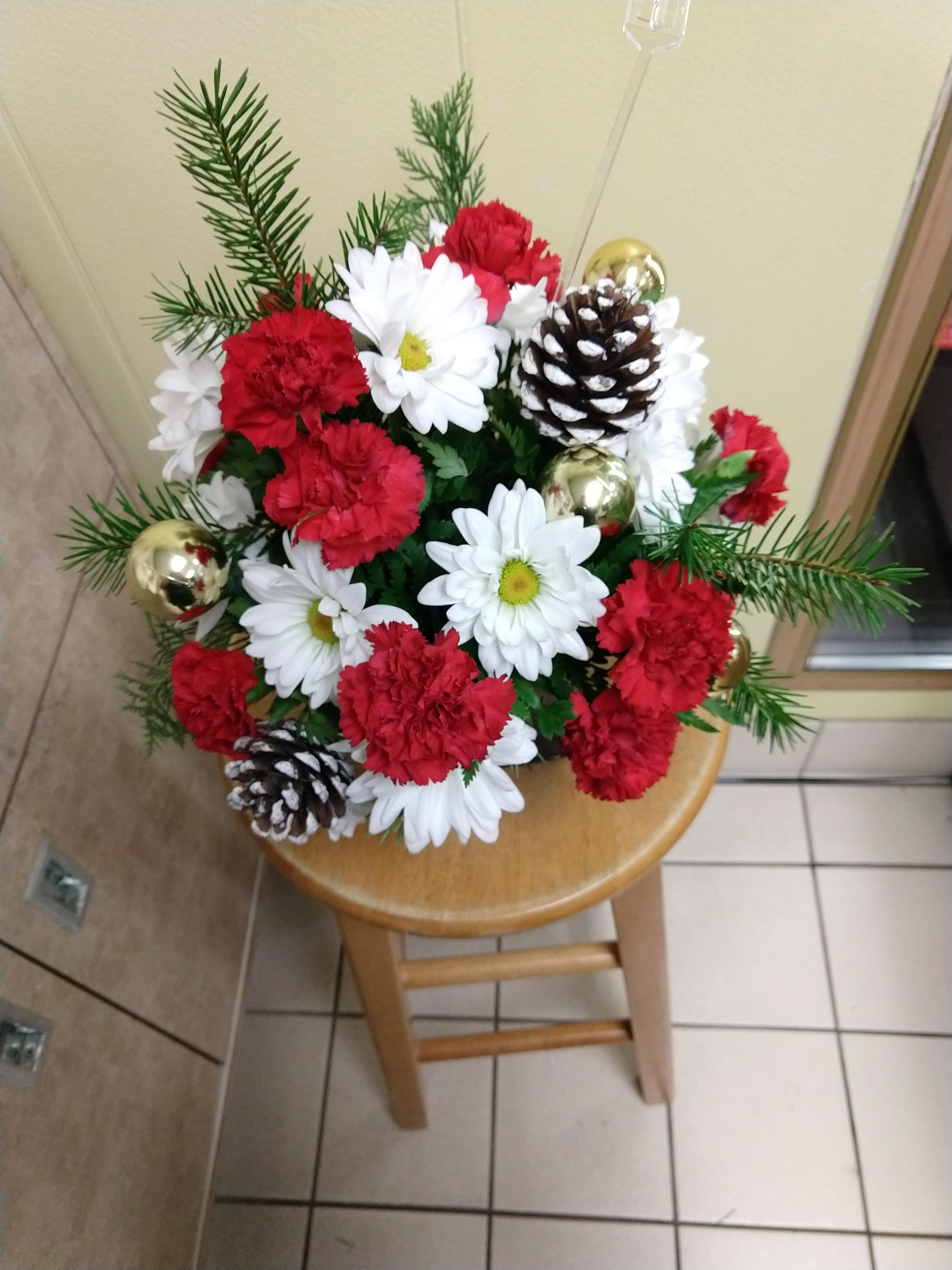 A winter arrangement filled with red carnations, white daisy poms, Christmas greenery, white tipped pinecones, and gold ornament balls.